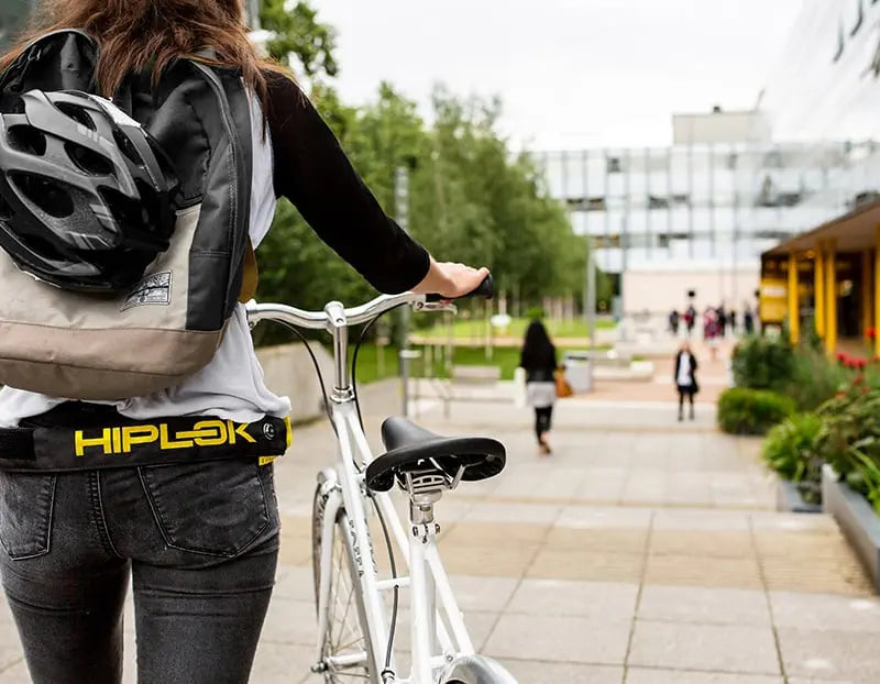 women walking bike at a univercity with a helmet strapped to rucksack and a bike chain lock strapped to her waist